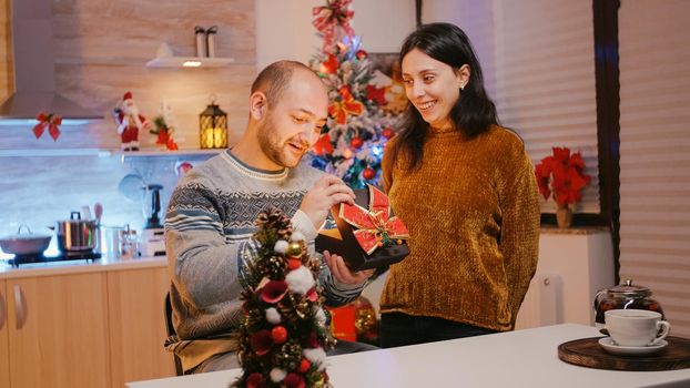 Woman giving gift box to man celebrating christmas eve. Adult receiving present with ribbon and wrapping paper from wife, enjoying holiday celebration. Couple in festive season