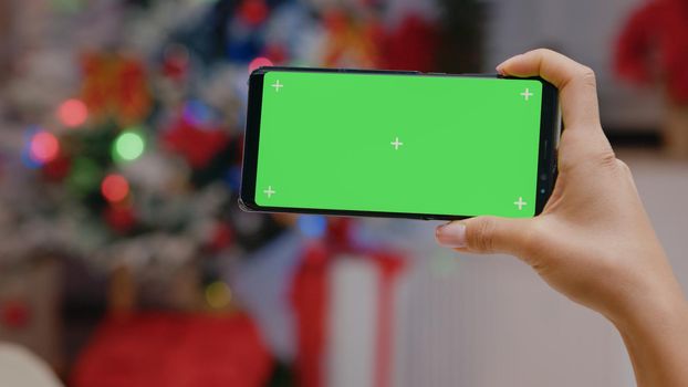 Close up of hand holding horizontal green screen on smartphone. Person looking at chroma key display with isolated template and mockup background in christmas decorated space