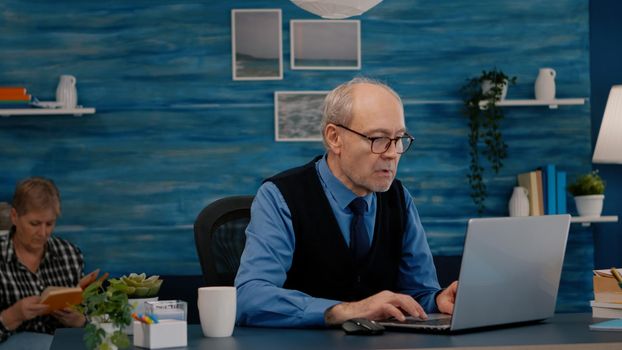 Old freelancer typing on laptop sitting in living room working remote. Busy focused elderly employee using modern technology reading typing, searching while old wife sitting on couch reading a book