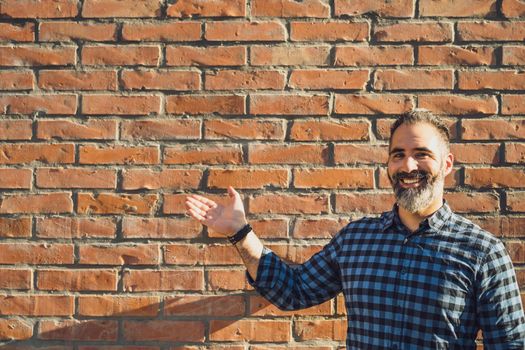 Portrait of modern businessman with beard gesturing while standing in front of brick wall outdoor.