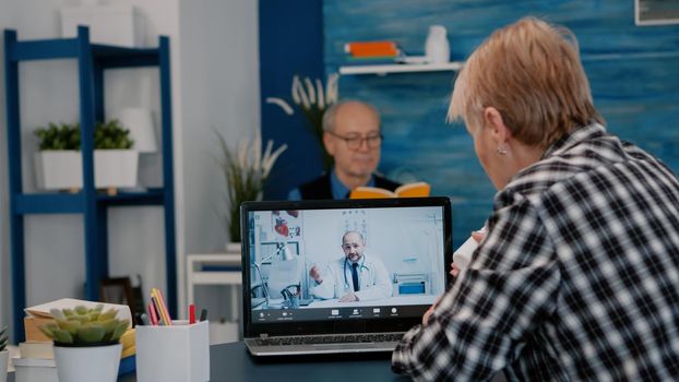 Young male doctor talking to senior female patient by tele medicine online video call in clinic. Physician using telehealth medical chat virtual healthcare visit on laptop screen, explaning treatment