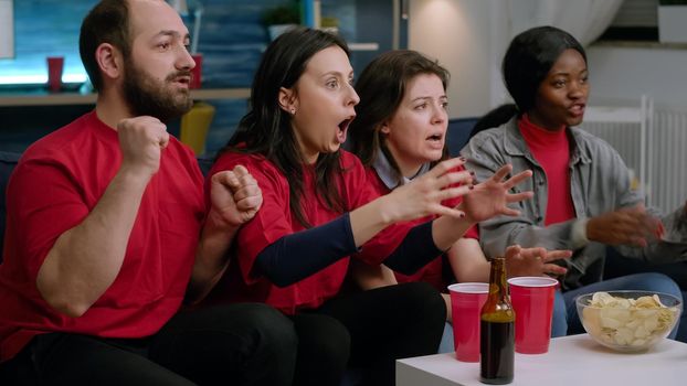 Group of mixed race sport fans watching football match on television while sitting on sofa late at night in living room. Multiethnic friends dissapointed after team losing game championship