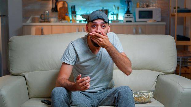 Concentrated man with eye sleep mask sitting in front of television while watching entertainment movie. Caucasian male with beard lying on sofa late at night in kitchen