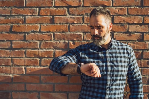 Modern businessman with beard looking at his watch while standing in front of brick wall outdoor.
