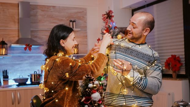 Festive couple trying to untangle garland of twinkle lights while decorating kitchen. Man and woman feeling cheerful and getting tangled in string of christmas lights with illuminated bulbs