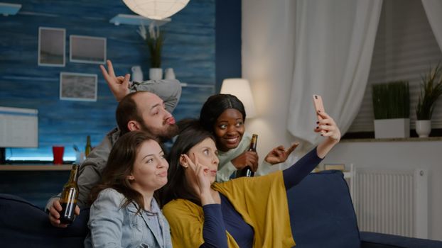 Multiracial friends resting on sofa enjoy spending time together while taking selfie sharing on social media. Group of multi-ethnic people laughing late at night in living room
