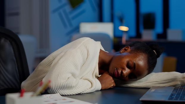 Dark skin freelancer doing overtime falling asleep with hand on desk trying to respect deadline for project. African employee working late at financial project sleeping on table in strat up office