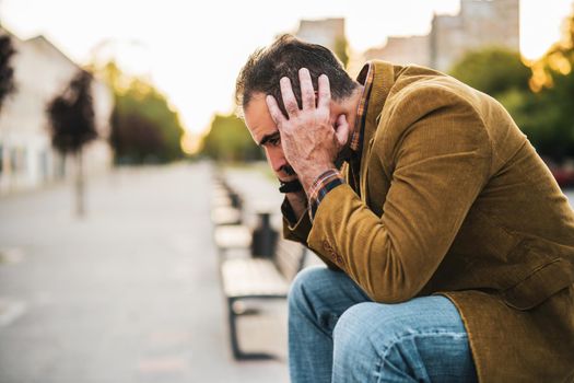 Depressed businessman having headache and sitting on a bench in the city street.