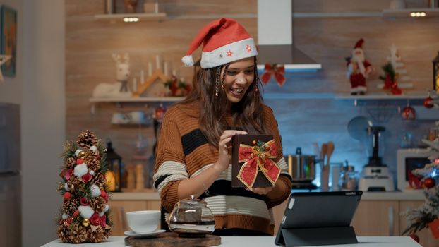 Smiling woman using video call technology for presents in festive decorated kitchen with tree and ornaments. Person with santa hat giving virtual gifts to family on christmas eve day