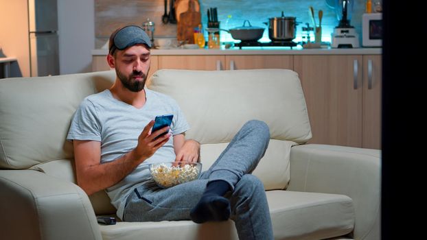 Bored man sitting on couch holding popcorn bowl while texting message on social network using modern phone. Caucasian male watching tv sports series late at night in kitchen