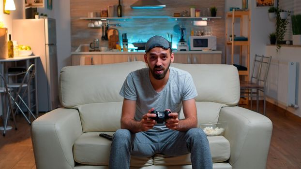 Upset pro gamer sitting in front of television losing soccer video games holding wireless controller. Frustrated man with sleep mask lying on sofa late at night in kitchen