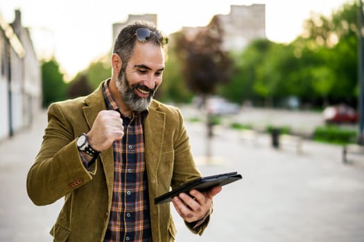 Excited modern businessman with beard using digital tablet while standing in on the city street.