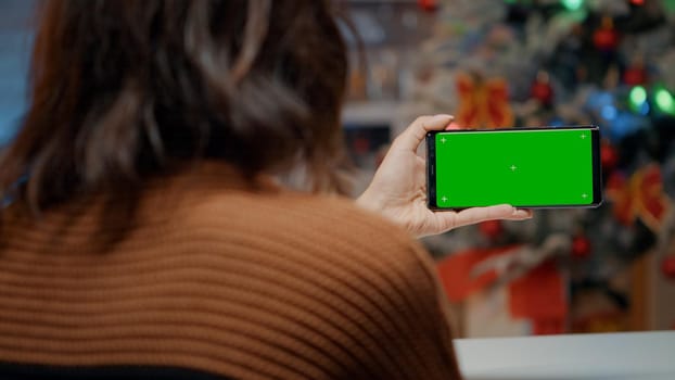 Young woman using smartphone in festive kitchen decorated with ornaments and tree. Adult holding green screen background device for digital seasonal holiday celebration dinner at home