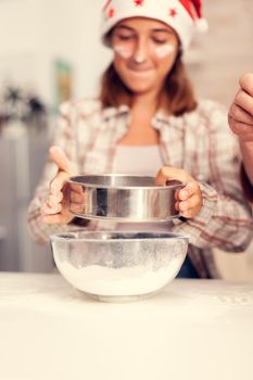 Joyfull child making cookies on christmas day sifting flour through sieve. Happy cheerful teenage girl helping senior woman preparing sweet cookies to celebrate winter holidays wearing santa hat.