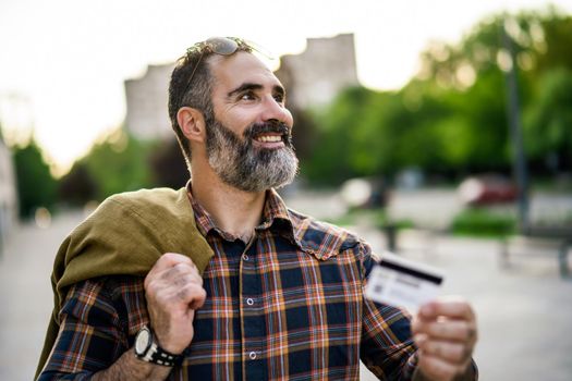 Portrait of modern businessman with beard holding credit card while standing on the city street.