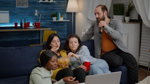 Multiracial friends chilling on sofa in living room during movie night watching online comedy film using laptop computer. Group of mixed people hanging out together, drinking beer, eating snacks