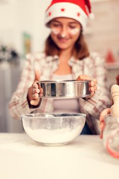 Girl sifts flour through a sieve on christmas day for baking cookies. Happy cheerful joyfull teenage girl helping senior woman preparing sweet biscuits to celebrate winter holidays wearing santa hat.
