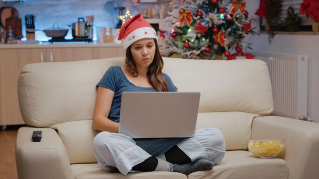 Bored adult using laptop and looking at movie on television for christmas eve celebration. Woman wearing santa hat with device, watching TV and feeling lonely on holiday festivity.