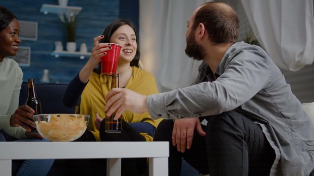 Multiracial friends laughing while enjoying night party drinking beer, sharing funny story relaxing on sofa late in living room. Group of mixed race people hanging out spending time together