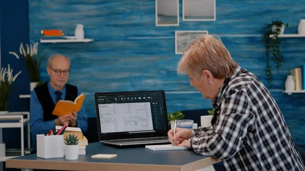 Senior woman architect analysing digital prototype with plans from laptop working from home, taking notes on notebook. Elderly designer using cad software to create a 3D concept of buildings