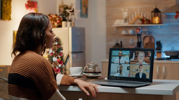 Young adult using laptop for work video call in kitchen decorated with tree and ornaments preparing for holiday dinner celebration on christmas day. Woman using virtual communication at home