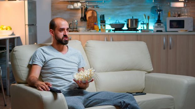 Upset caucasian man watching sport match alone in the living room while hus favourite team looses the game, eating popcorn late at night