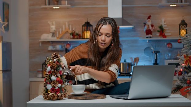 Festive woman talking on video call using laptop in christmas decorated room. Young adult celebrating traditional holiday on internet comunication at home pouring cup of tea from kettle