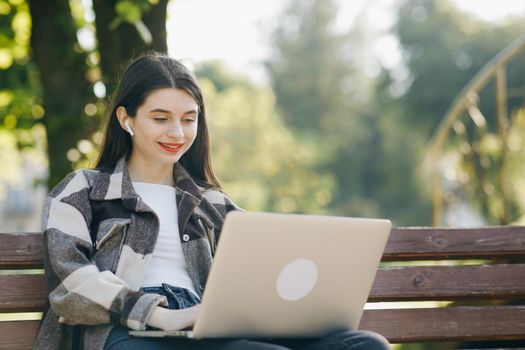 Caucasian woman in headset typing on the laptop computer on the bench in the park. Female student makes conference video call on laptop computer talks with web tutor, online teacher in webcam chat.