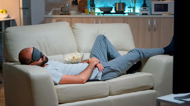 Man eye sleep mask falling asleep while watching movie, sitting in front of television. Caucasian male with beard looking entertainment series late at night in kitchen