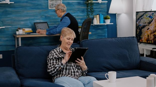 Happy old senior grandmother holding digital tablet browsing, searching informations, shopping online, while adult man working at laptop in background. Person enjoying using notepad gadget