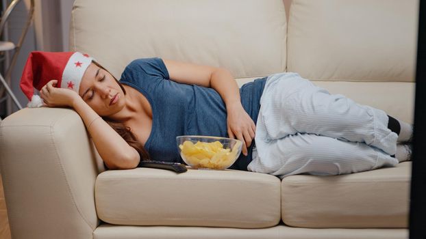 Close up of woman sleeping on couch at television while having santa hat on head for christmas eve celebration. Adult laying on sofa falling asleep with bowl of chips on holiday