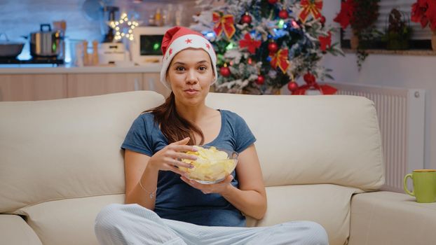 POV of woman using video call communication and eating chips. Festive adult talking to family on online remote conference for christmas eve festivity. Cheerful person looking at camera