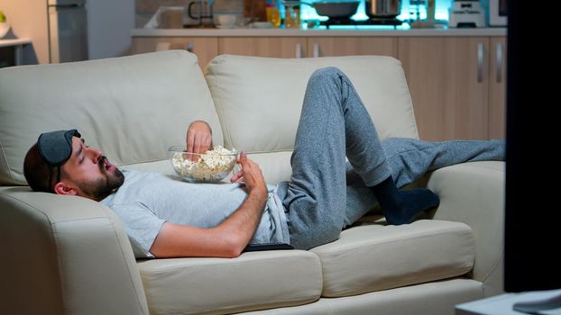 Caucasian man lying in sofa, eating popcron and watching TV late at night in the living room. Enjoying midnight snack, television entertainment, leisure lifestyle evening show
