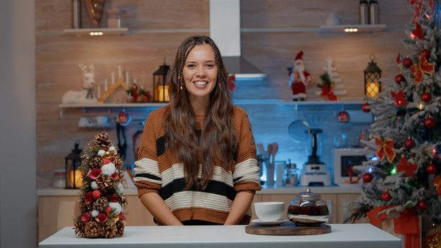 Woman talking on video call in decorated kitchen at home for christmas festivity in december. Caucasian young adult chatting online on internet with friends while drinking tea from hot pot