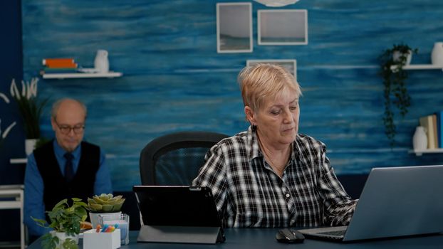 Multitasking retired woman reading on laptop and tablet in same time working from home. Focused old remote employee searching, analysing financial reports while elderly husband reading a book