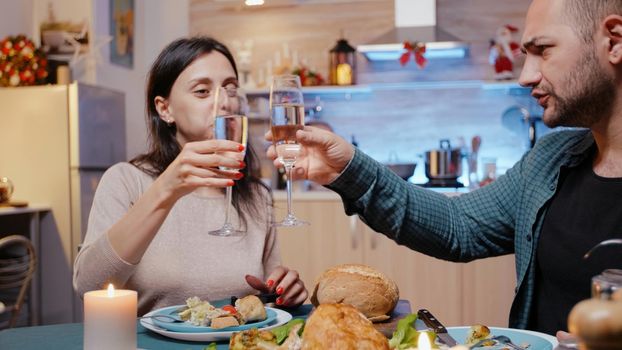 Man and woman eating chicken and clinking glasses with champagne at festive dinner. Couple celebrating christmas eve with traditional meal and alcohol on table. People enjoying festivity