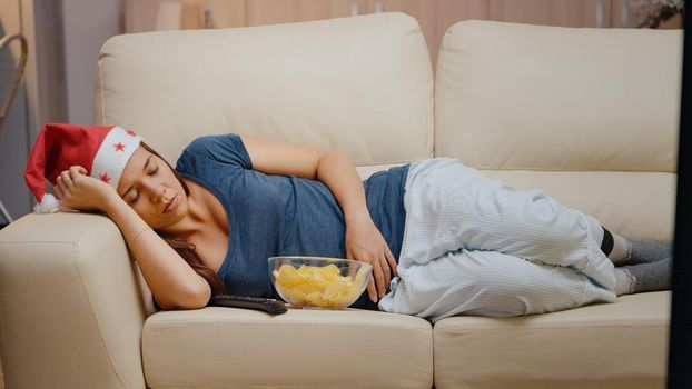 Close up of tired woman with santa hat sleeping on sofa at television. Festive person falling asleep on couch while watching TV, having remote control and bowl of chips on christmas eve.