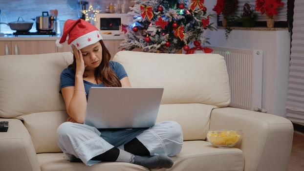 Tired woman falling asleep while working on laptop on christmas eve. Festive person with santa hat feeling sleepy and sitting on couch with device for winter holiday celebration.