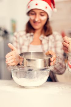 Grandchild on christmas day sifting flour for cookies. Happy cheerful joyfull teenage girl helping senior woman preparing sweet cookies to celebrate winter holidays wearing santa hat.