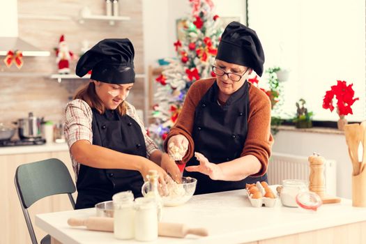 Kid wearing apron mixing dough on christmas day with grandmother. Happy cheerful joyfull teenage girl wearing apron and bonette helping senior woman preparing sweet cookies to celebrate winter holidays.