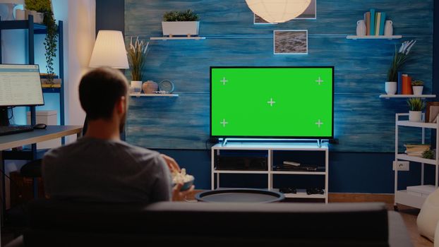 Caucasian person looking at green screen tv in living room. Man watching chroma key blank display using mockup template background in cozy living room eating pop corn late at night