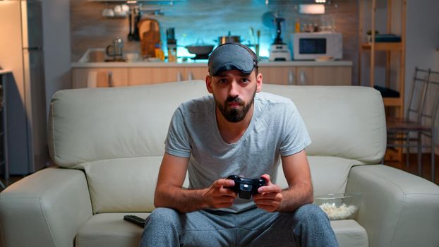 Front view of pro gamer sitting on sofa in front of television while playing videogames competition using wireless joystick. Cheerful man with sleep mask enjoying time alone late at night in kitchen