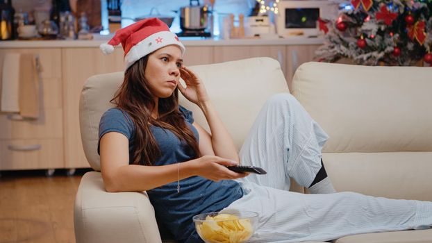 Frustrated woman using TV remote control to switch channels and wearing santa hat. Bored adult watching television and eating chips from bowl on christmas eve. Person on holiday