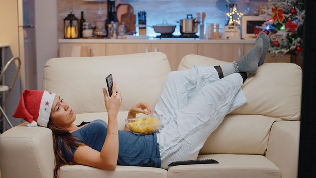 Woman browsing internet on smartphone and watching television on christmas eve. Festive adult looking at TV and phone screen for fun while eating chips from bowl. Person with santa hat