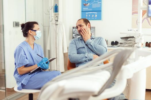 Dental assistant writing stomatology treatment on clipboard in dental clinic during sick patient check up. Woman nurse talking with man about teeth problem waiting medical examination