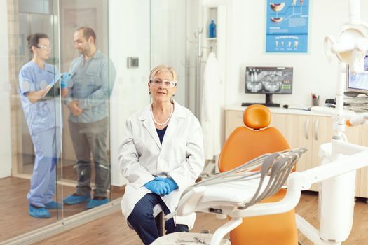 Portrait of senior dentist woman sitting on chair in stomatological hospital office. In background dentist assistant talking with sick patient for dental treatment during stomatology consultation