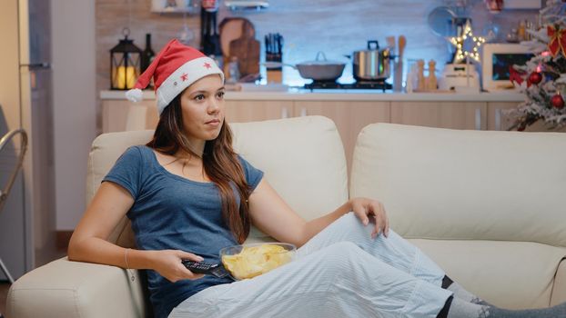 Cheerful adult wearing santa hat and watching television with snack on christmas eve. Festive woman looking at TV and switching channels while holding remote control in decorated space.
