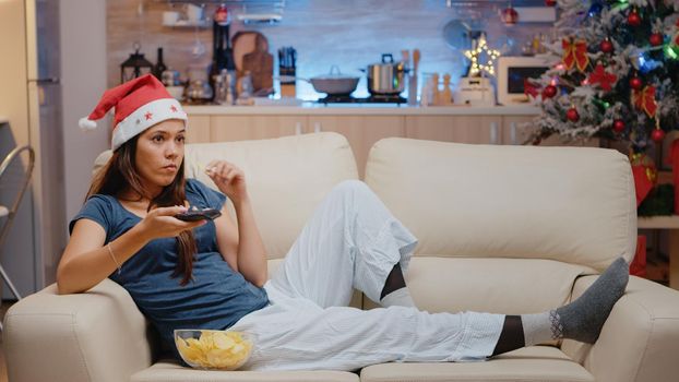 Bored woman switching channels and wearing santa hat on couch in festive space. Adult watching television and using TV remote control on christmas eve. Person with technology on holiday