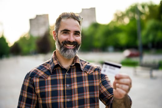 Portrait of modern businessman with beard holding credit card while standing on the city street.