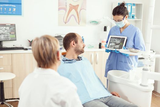Stomatologist nurse in gloves holding tablet showing jaw x-ray to sick patient while discussing toothache treatment during dental consultation. Orthodontist talking to man in somatology office